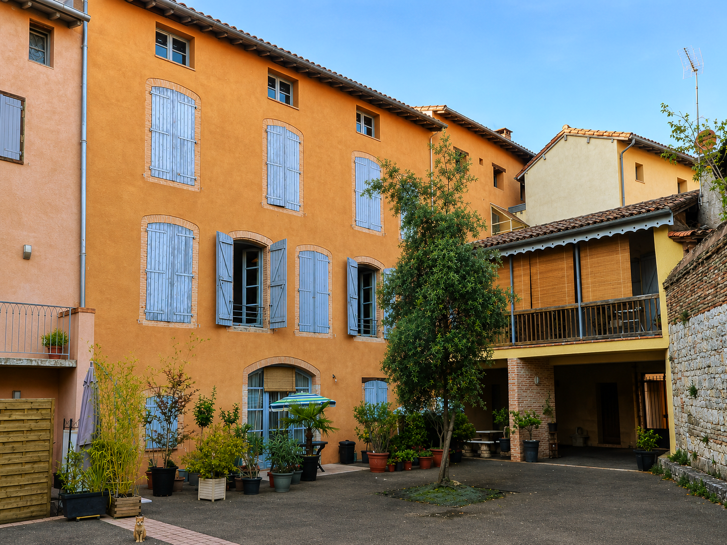 Bâtiment jaune de trois étages avec volets bleus, cour et petit arbre.