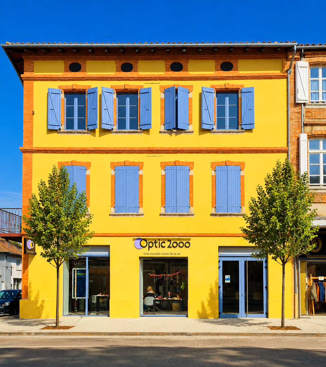 Bâtiment de trois étages avec volets bleus et enseigne Optic 2000 ; façade en briques jaunes.