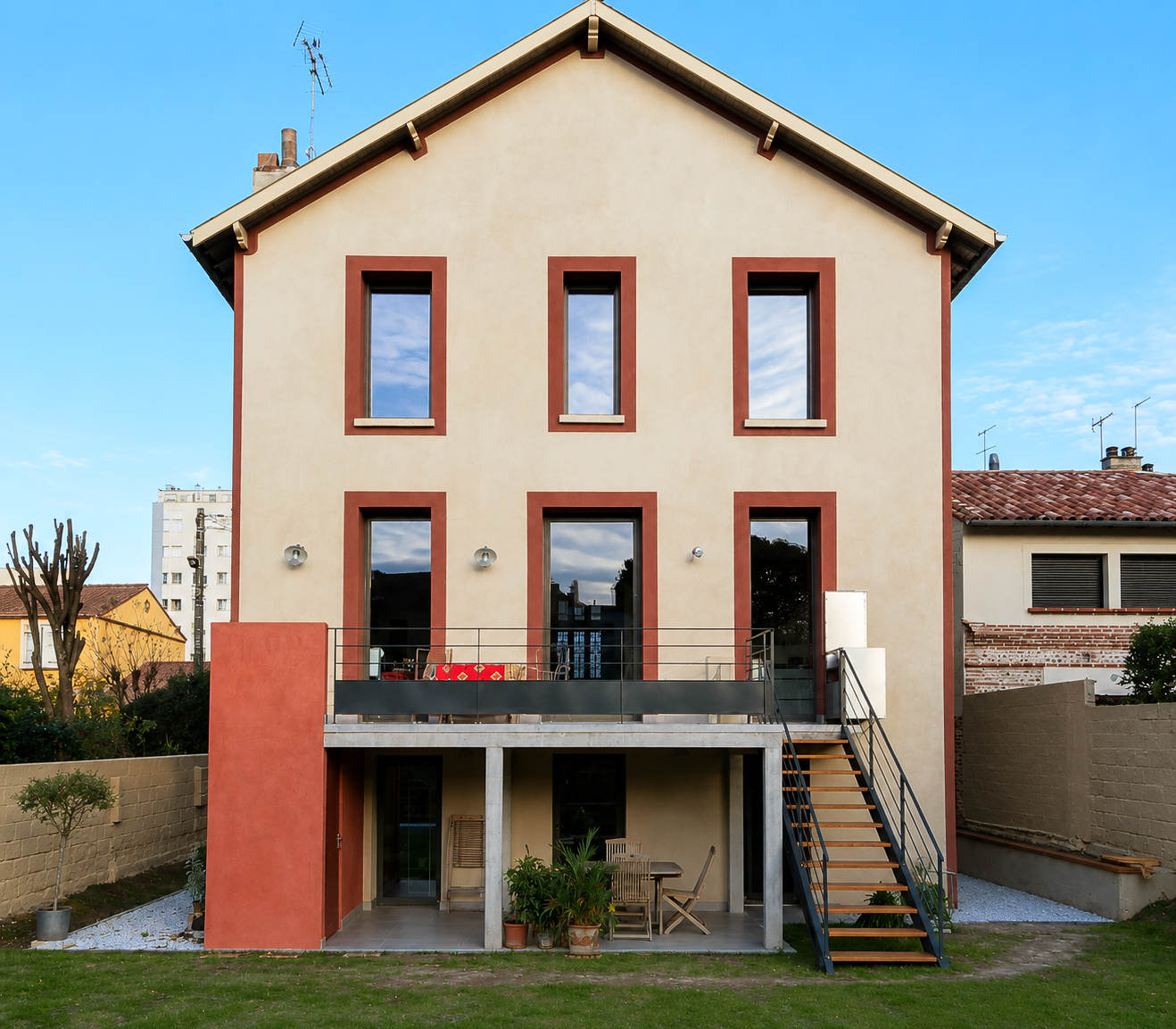 Maison beige avec six fenêtres, boiseries marron, petit balcon et escalier latéral.
