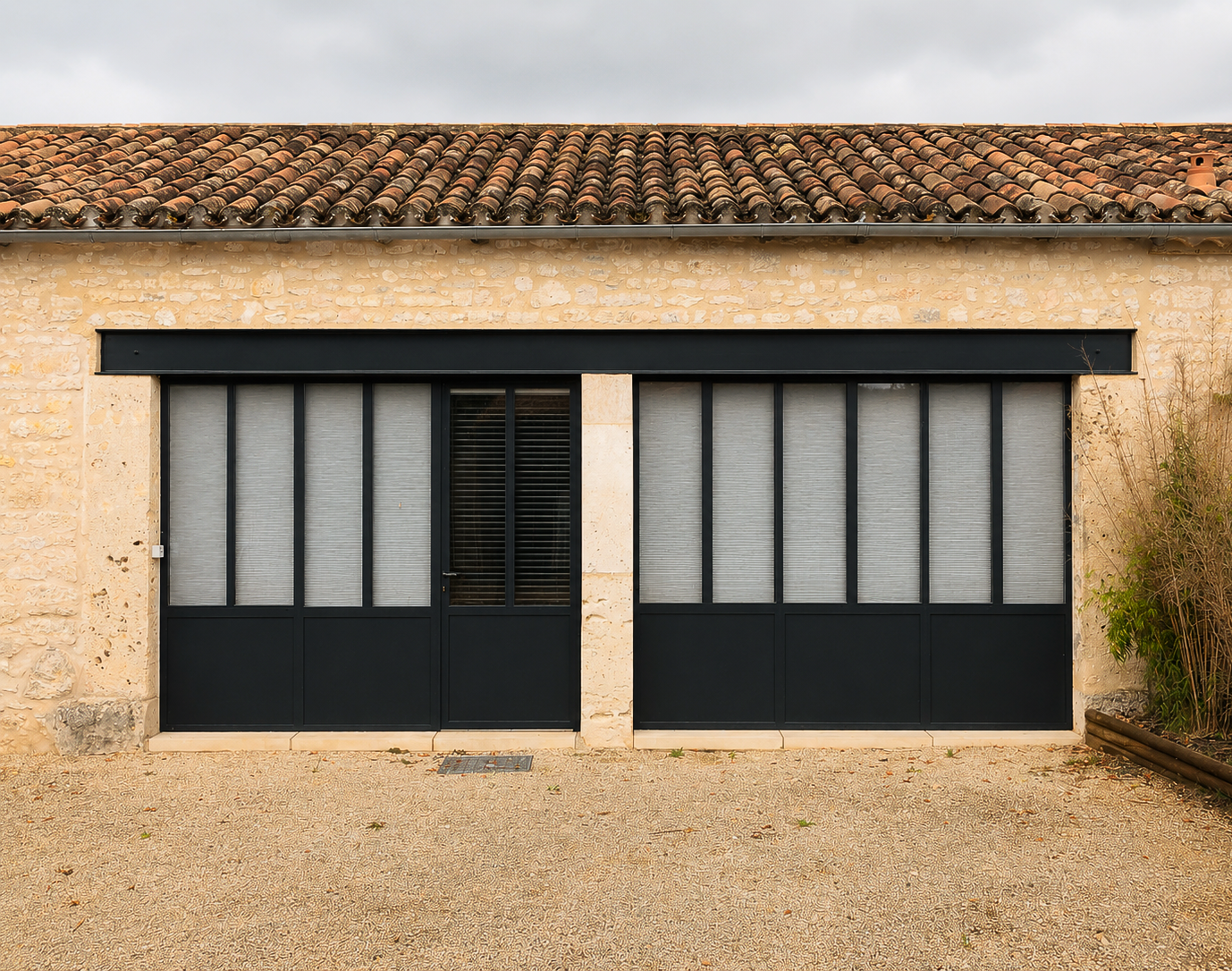 Une cour gravillonnée avec des bâtiments ; une porte-fenêtre à cadre sombre, un toit de tuiles rouges et une grande plante verte.