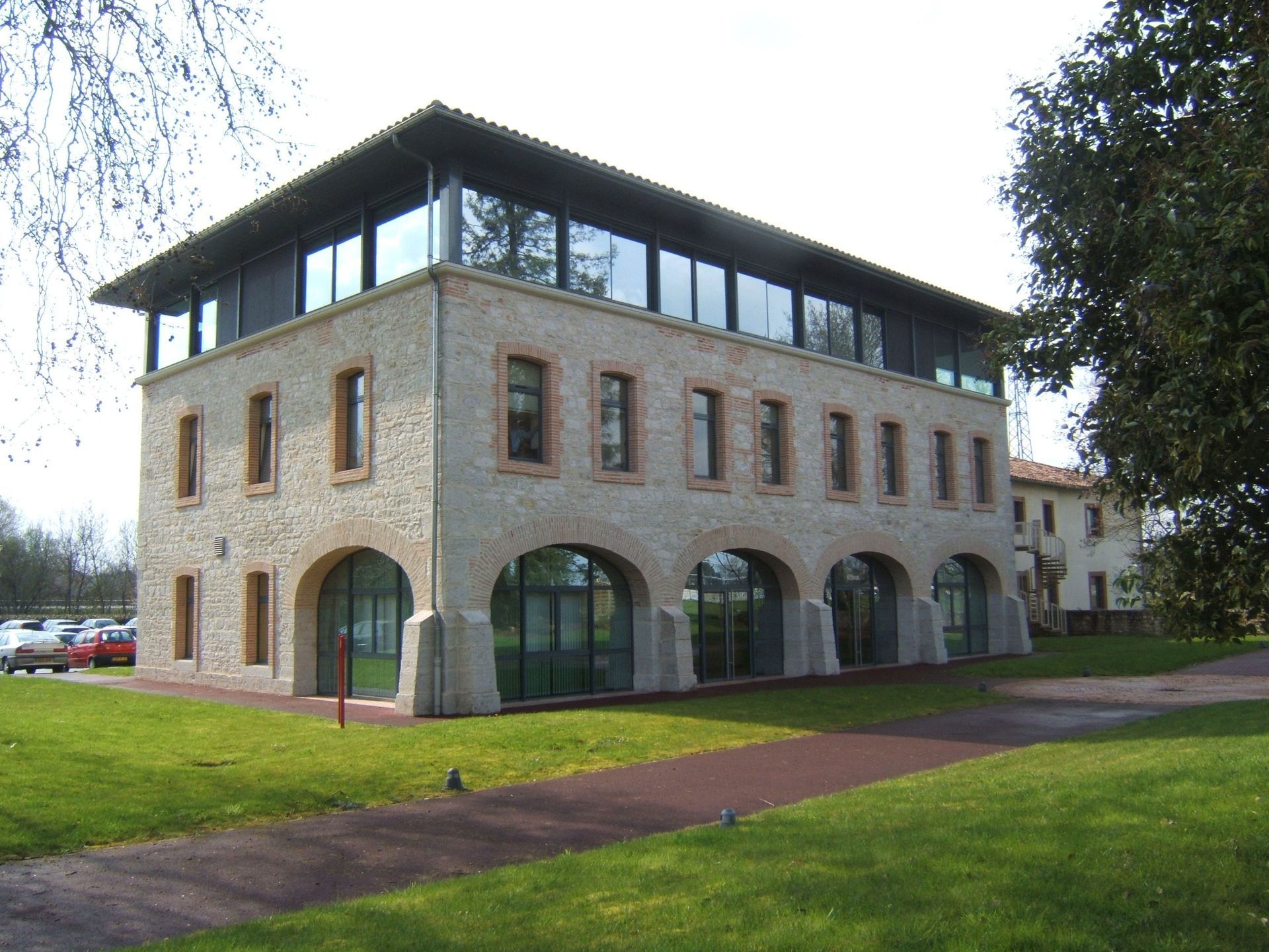 Bâtiment en pierre avec un étage supérieur vitré, des fenêtres cintrées au rez-de-chaussée, une pelouse verdoyante et des arbres.