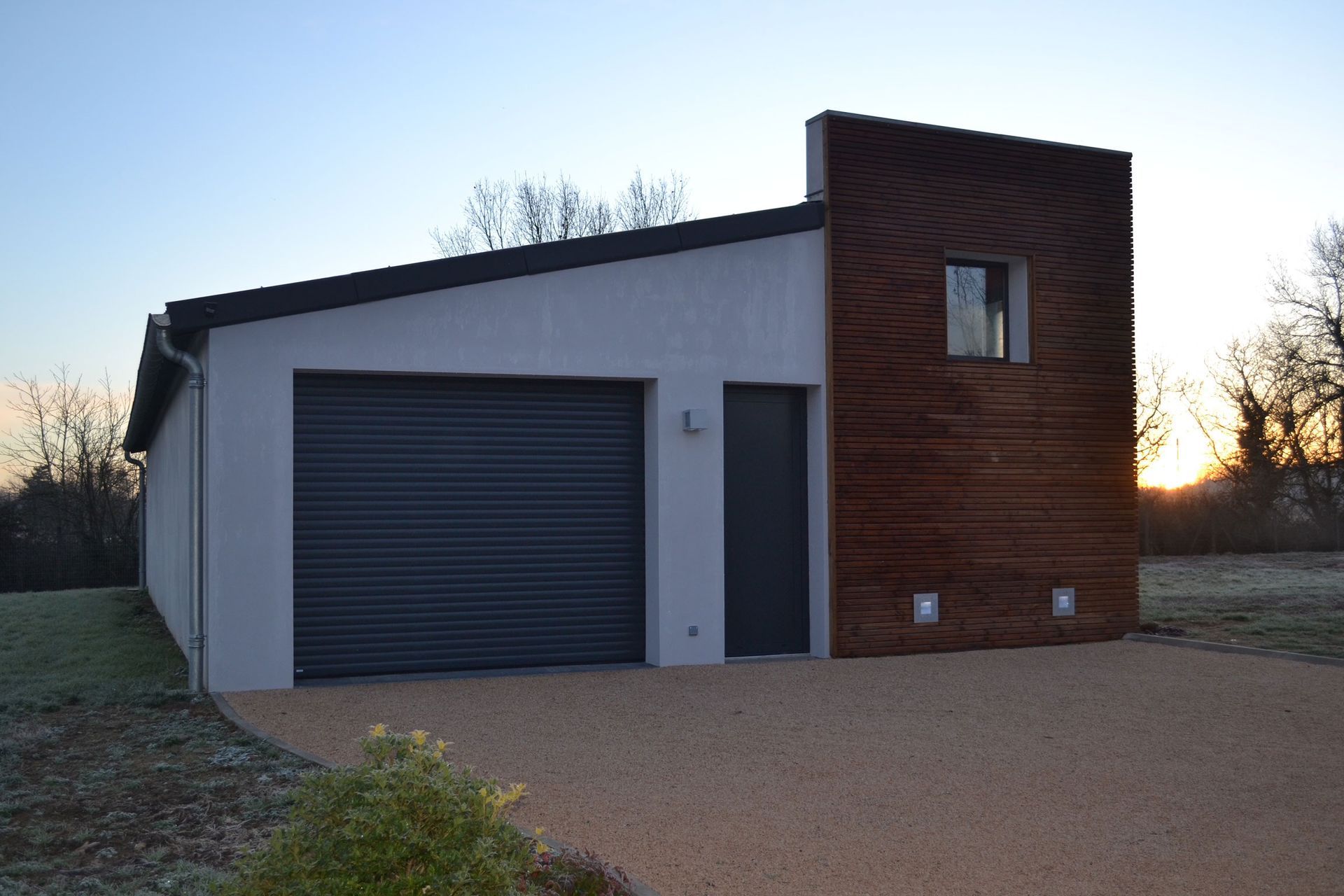 Garage avec porte grise, façade en bois foncé et allée de gravier sous un ciel dégagé.
