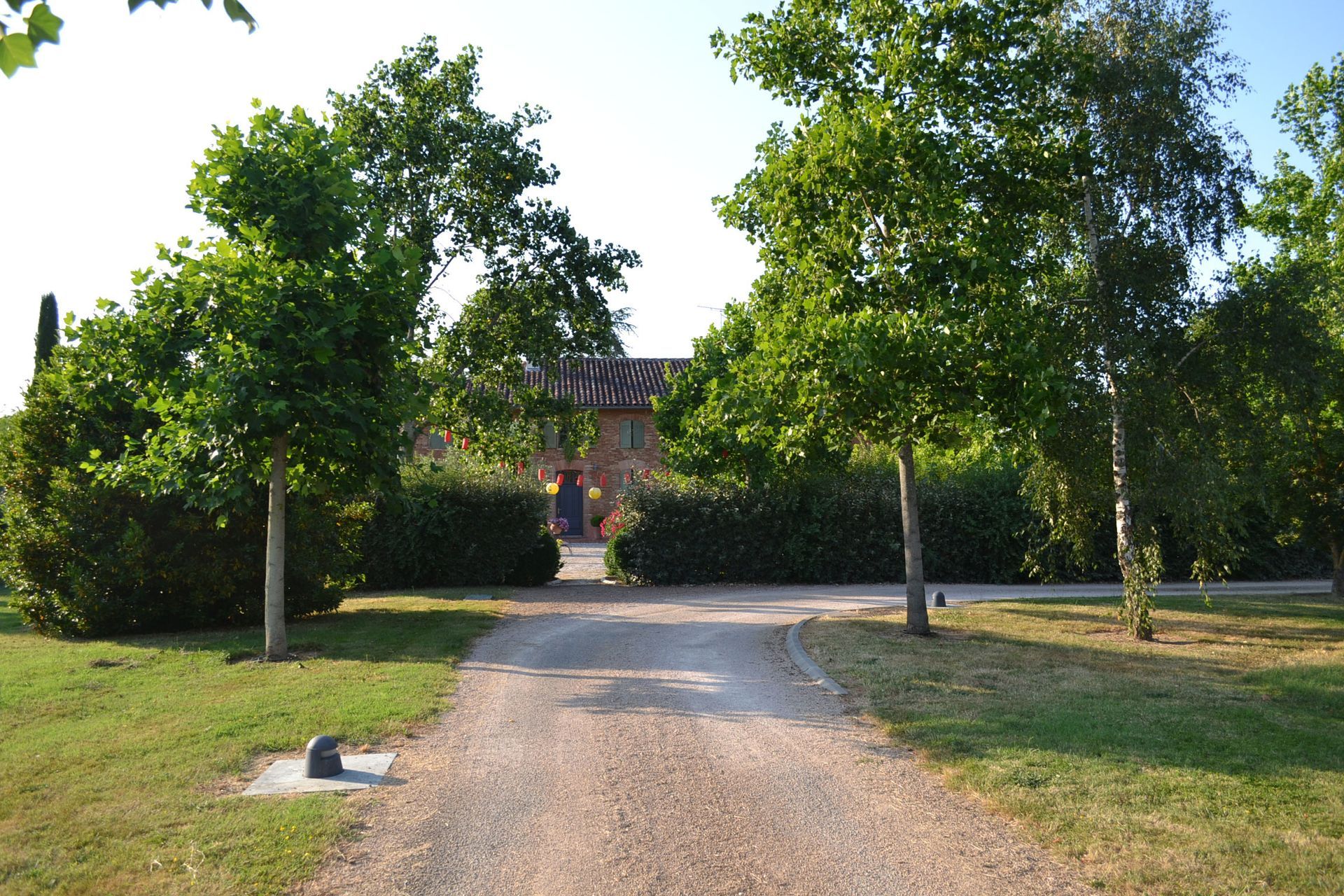 Chemin de gravier menant à un bâtiment encadré d'arbres et de buissons par une journée ensoleillée.