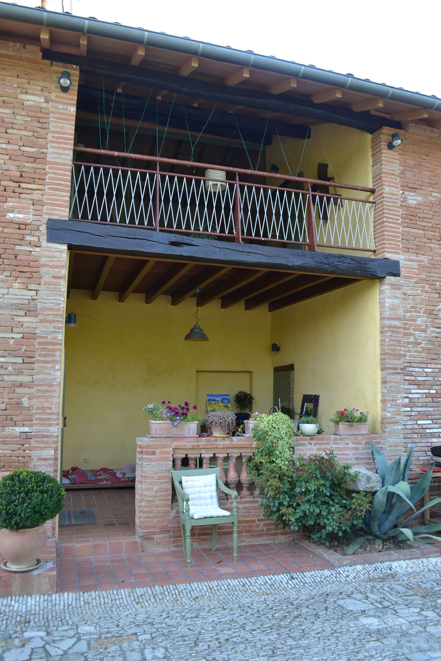 Immeuble en briques avec balcon au premier étage et patio au rez-de-chaussée. Chaise verte, plantes en pot et mur jaune.