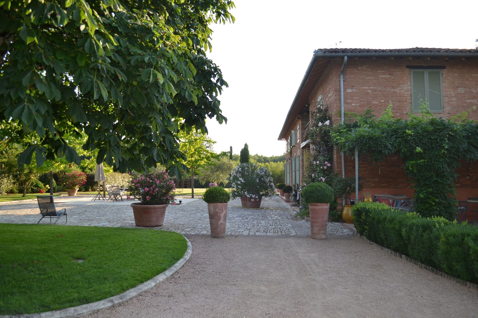 Un bâtiment en briques avec des plantes, une allée de gravier et une pelouse verdoyante dans un cadre extérieur ensoleillé.
