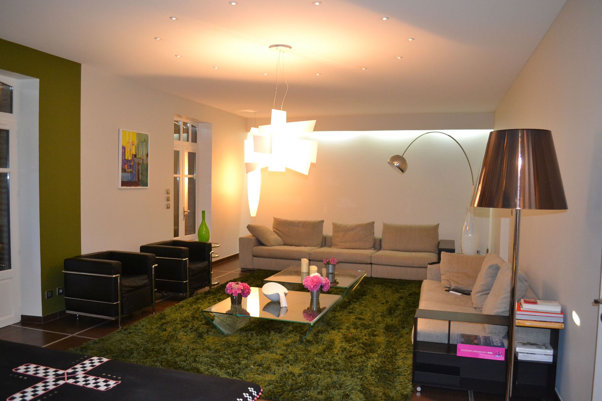 Salon avec tapis vert, canapé beige, fauteuils noirs et éclairage moderne.