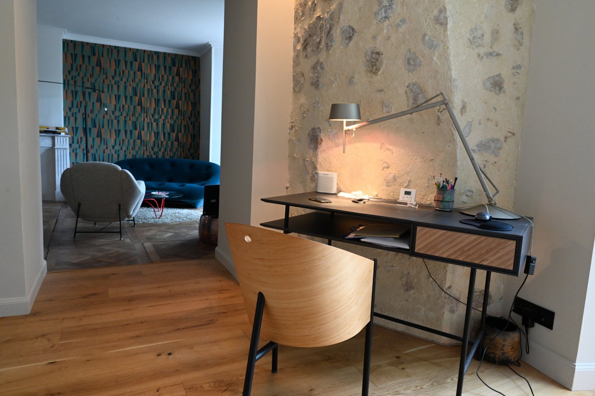 Bureau avec lampe et chaise, devant un mur en pierre. Vue intérieure d'une pièce avec un coin salon visible.