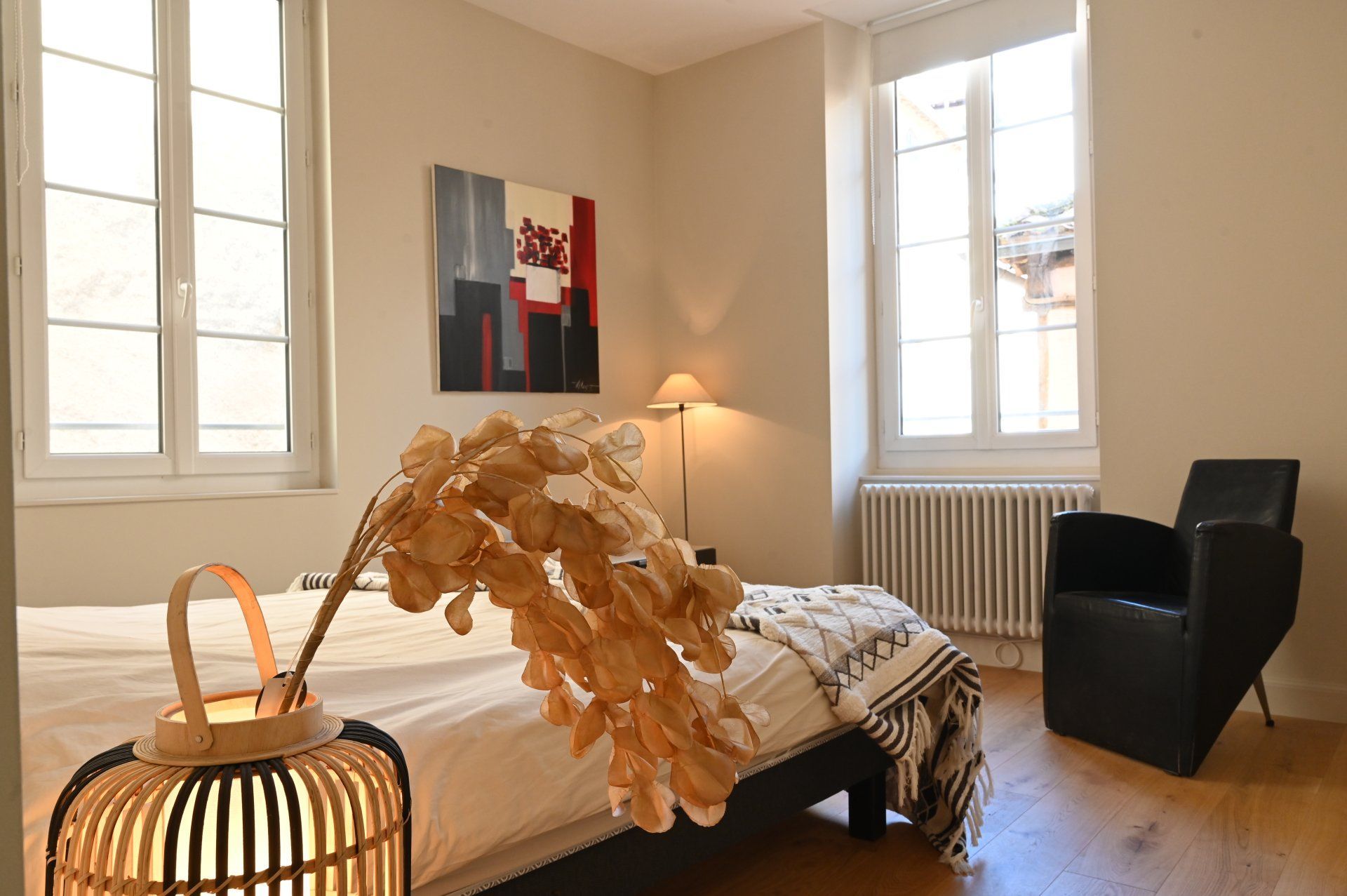 Chambre avec lit, fauteuil, œuvres d'art et fenêtres. Plante séchée décorative et lampe sur le lit.