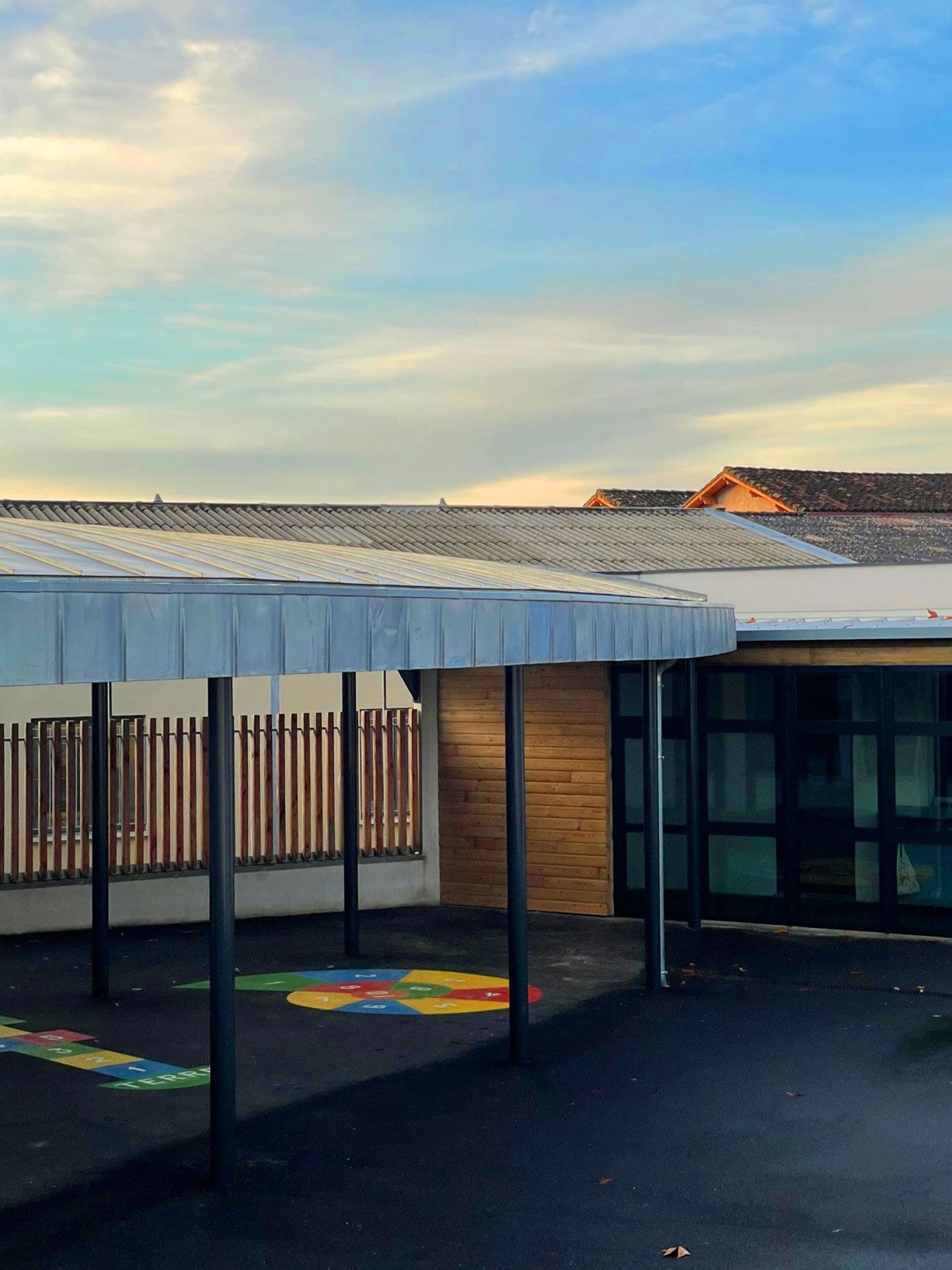 Extérieur du bâtiment scolaire avec allée couverte et aire de jeux colorée sous un ciel bleu.