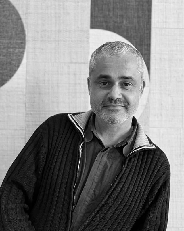 Homme aux cheveux clairs et à la barbe naissante, portant un pull foncé, souriant, appuyé contre un mur à motif géométrique.