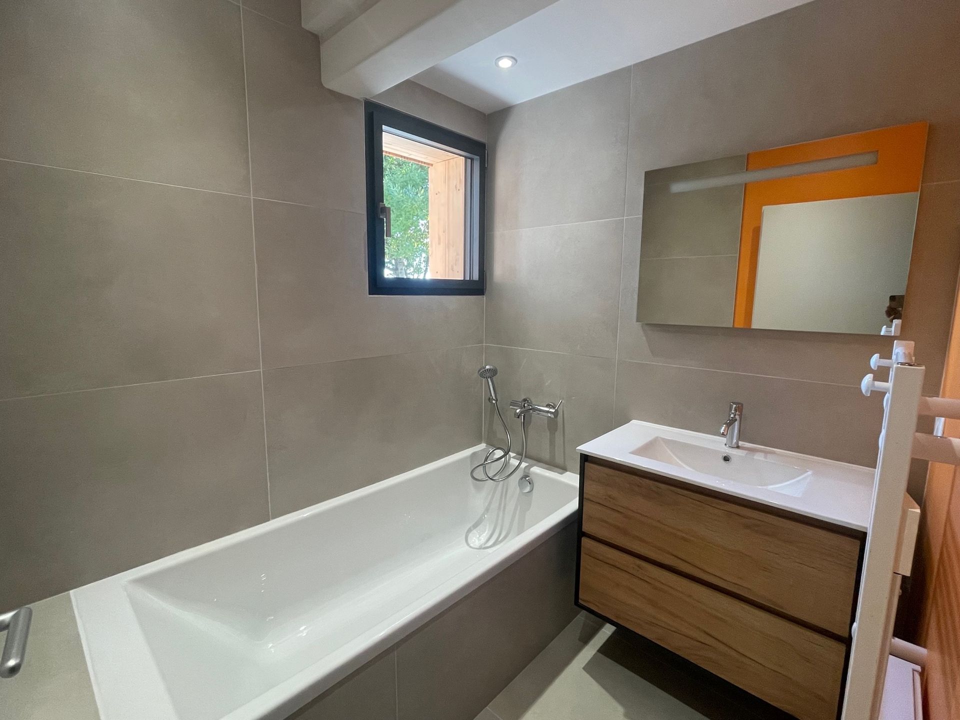 Salle de bains avec baignoire, meuble-lavabo en bois, murs carrelés gris et fenêtre.