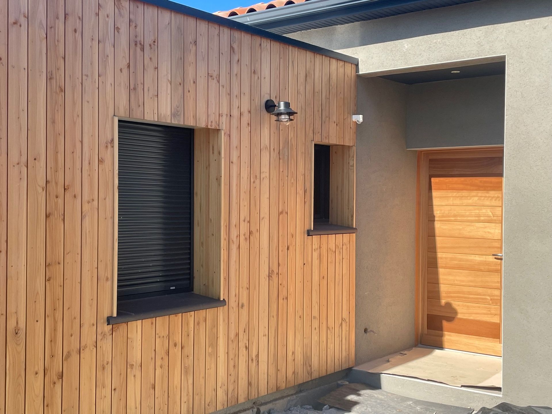 Mur extérieur en bois avec fenêtre, porte et luminaire ; palette de couleurs neutres.