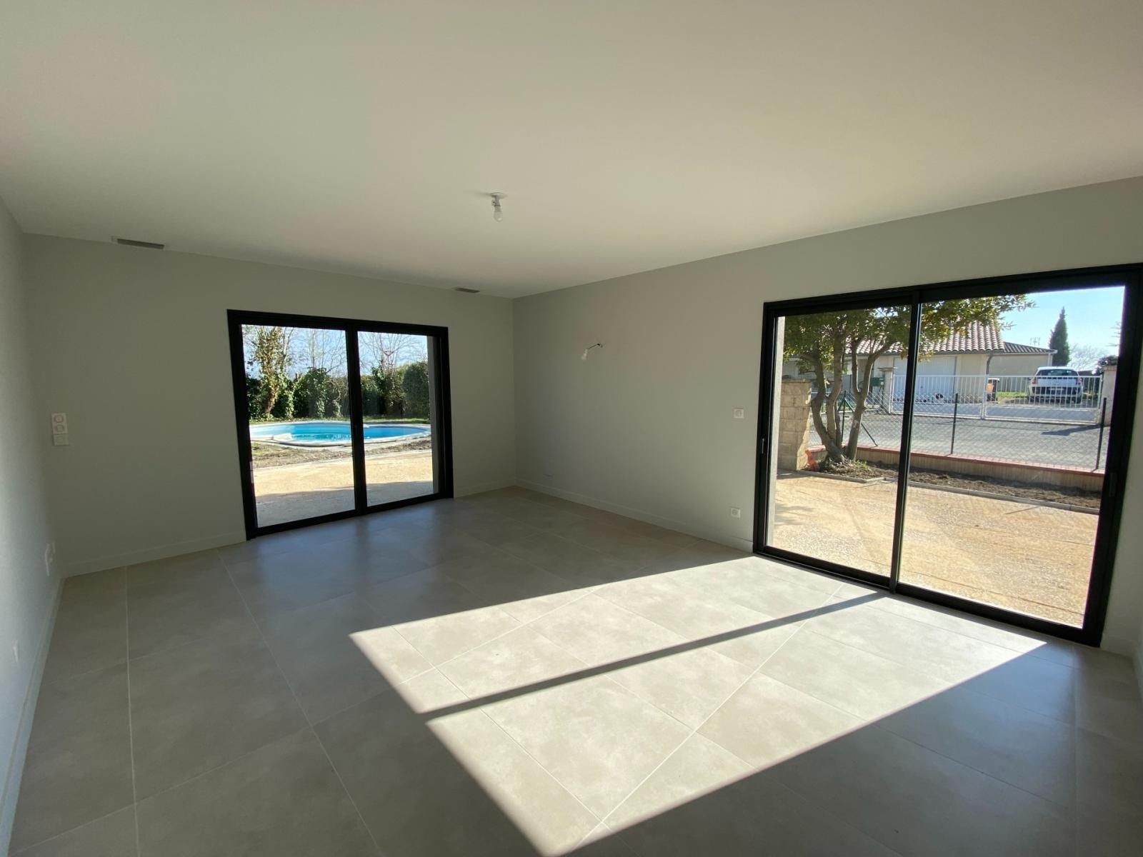 Pièce vide et ensoleillée, aux murs et au sol gris. Deux grandes portes coulissantes en verre offrent une vue sur l'extérieur.