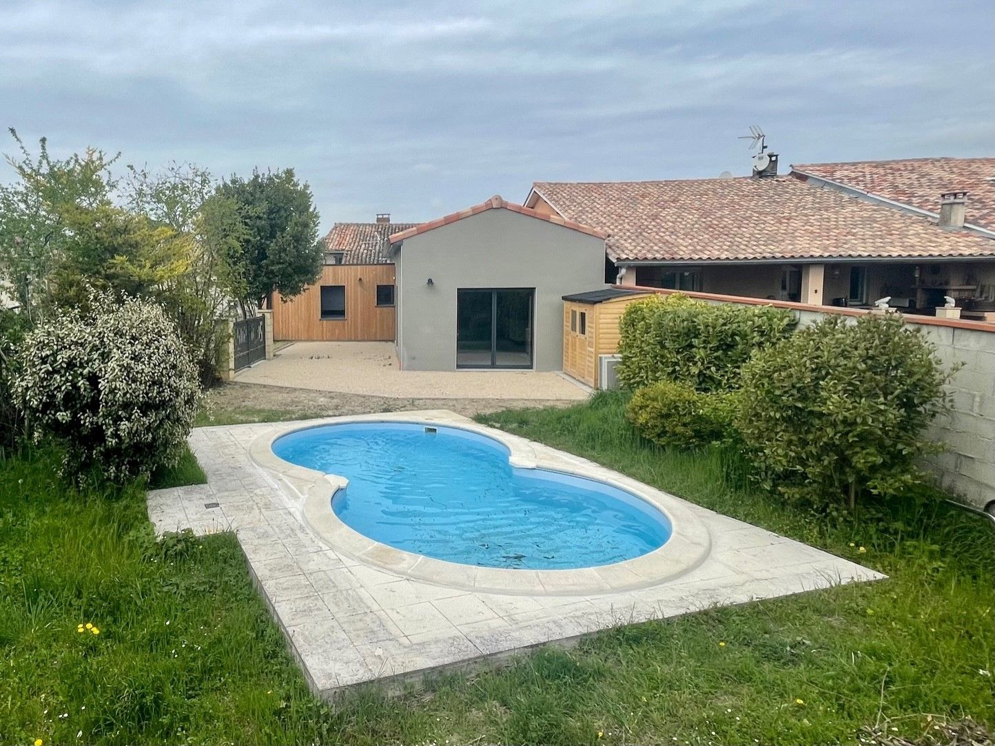Un petit jardin avec une piscine en forme de haricot. Un bâtiment gris se dresse à l'arrière-plan. Ciel couvert.