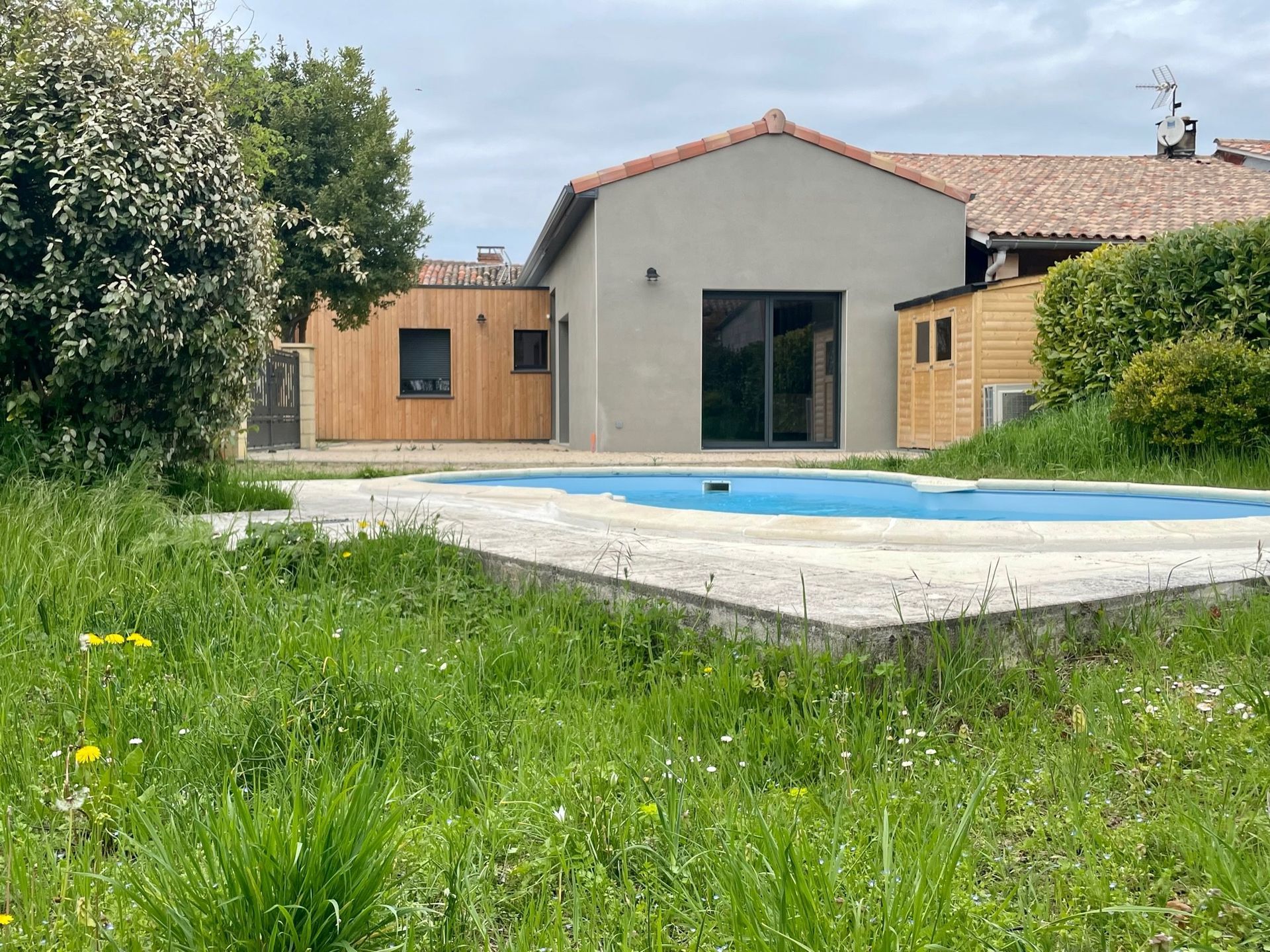 Jardin avec maison, piscine et abri de jardin. Pelouse verte, piscine bleue, bois clair, maison grise.
