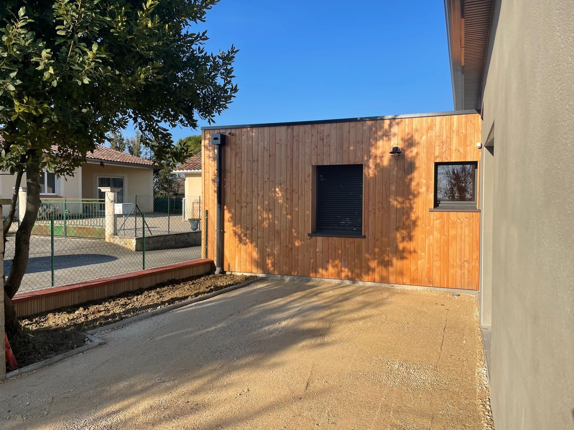 Bâtiment à façade en bois avec une petite fenêtre et une cour gravillonnée, jouxtant un mur en stuc, avec un arbre et des maisons en arrière-plan.