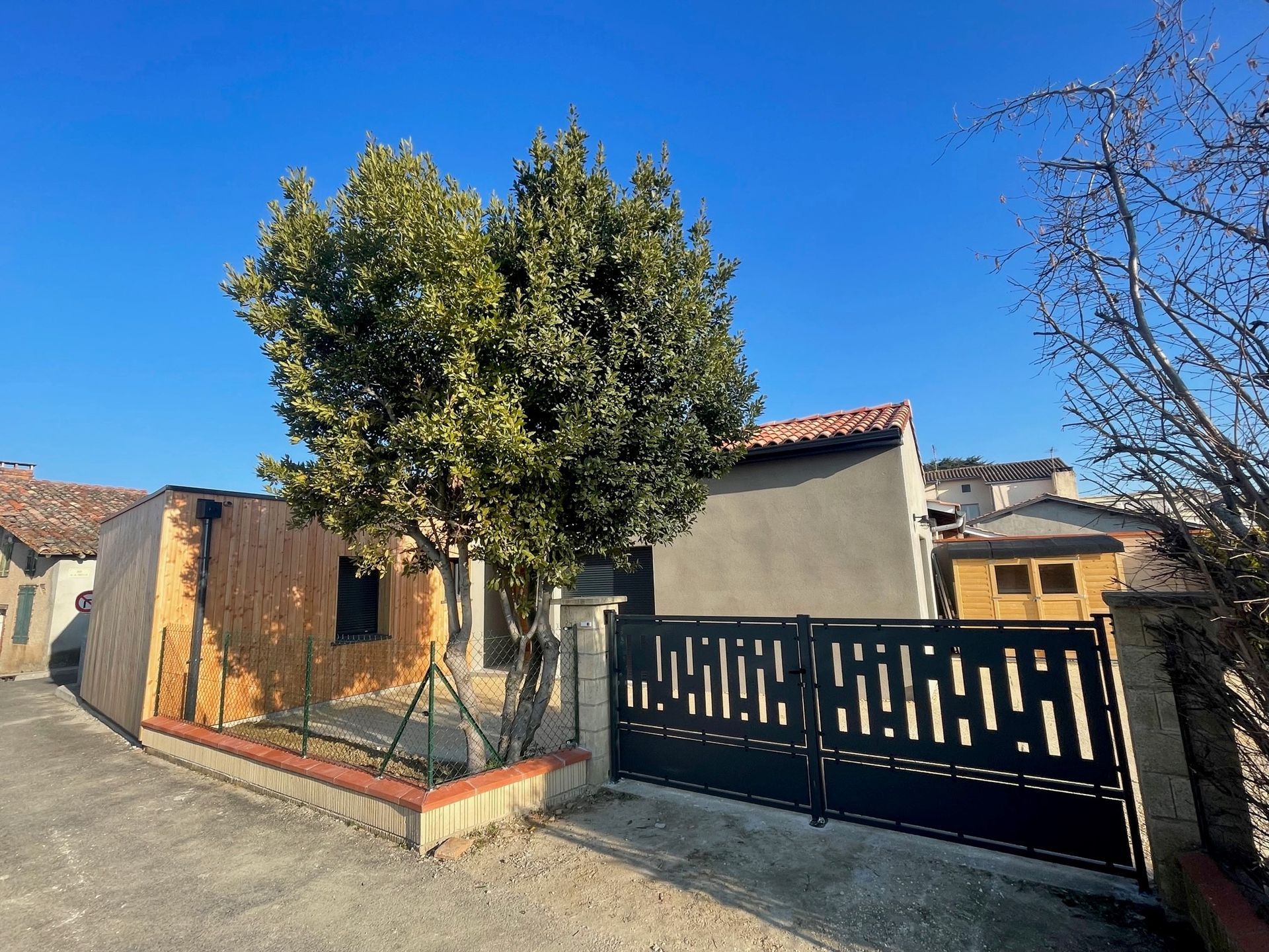 Extérieur d'une petite maison avec un portail en métal noir et un arbre devant, sous un ciel bleu.