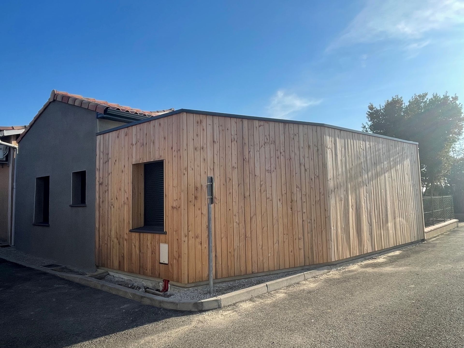 Extérieur d'un bâtiment à façade en stuc gris et en bois, sous un ciel bleu.