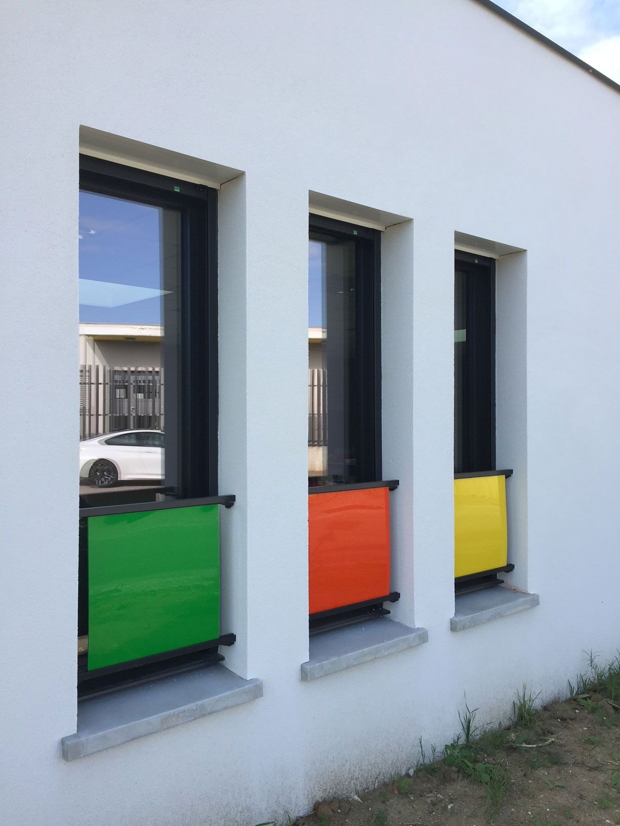 Trois fenêtres à panneaux colorés : vert, orange et jaune. Façade du bâtiment blanche.