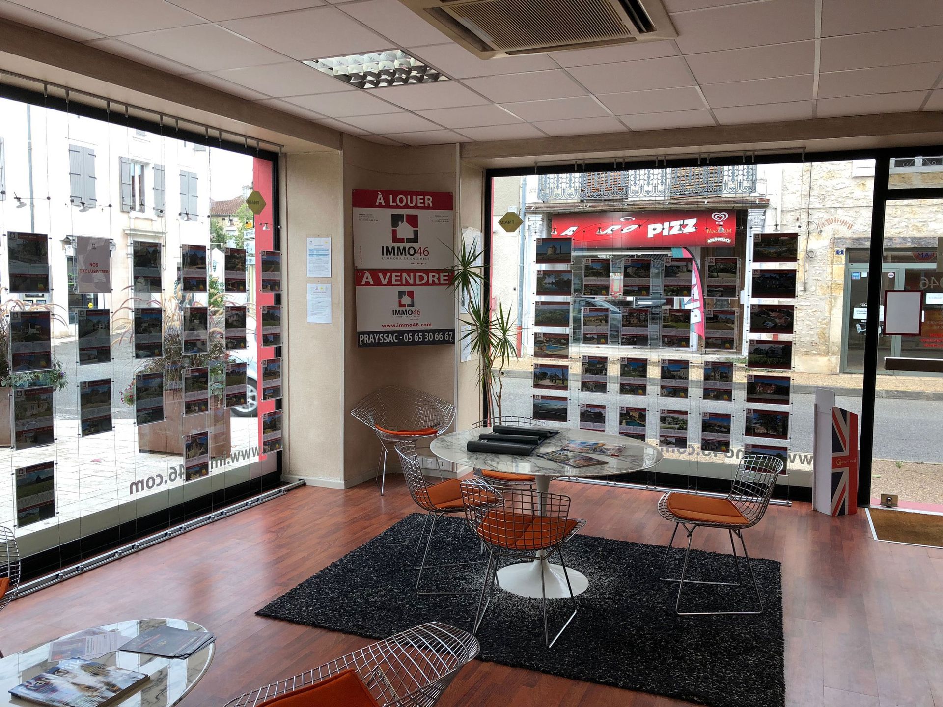 Intérieur d'une agence immobilière avec des annonces de biens immobiliers en vitrine et des sièges orange.