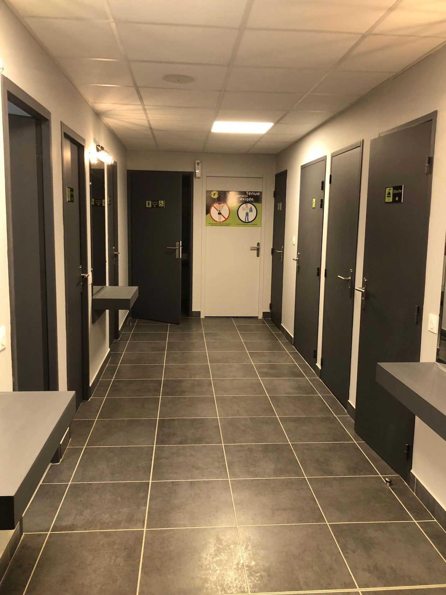 Couloir avec portes sombres, sol carrelé gris, bancs et éclairage au plafond.