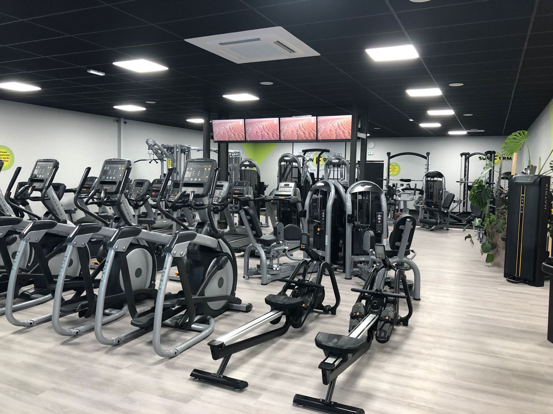 Intérieur de salle de sport avec divers appareils de musculation, notamment des vélos elliptiques et des rameurs.