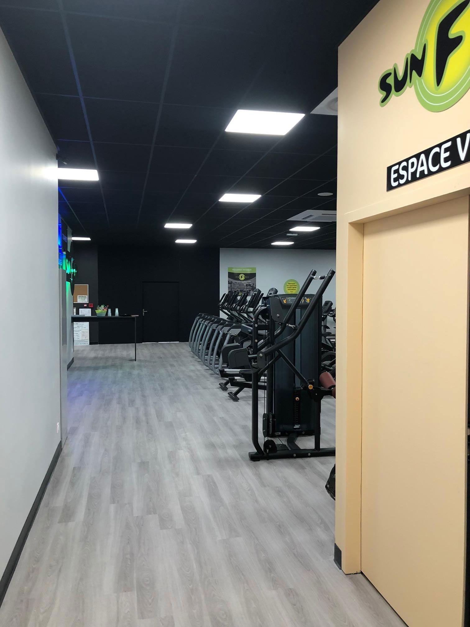 Un couloir de salle de sport avec des appareils de musculation, un sol gris et un plafond noir. Une pancarte indique SUN FUN.
