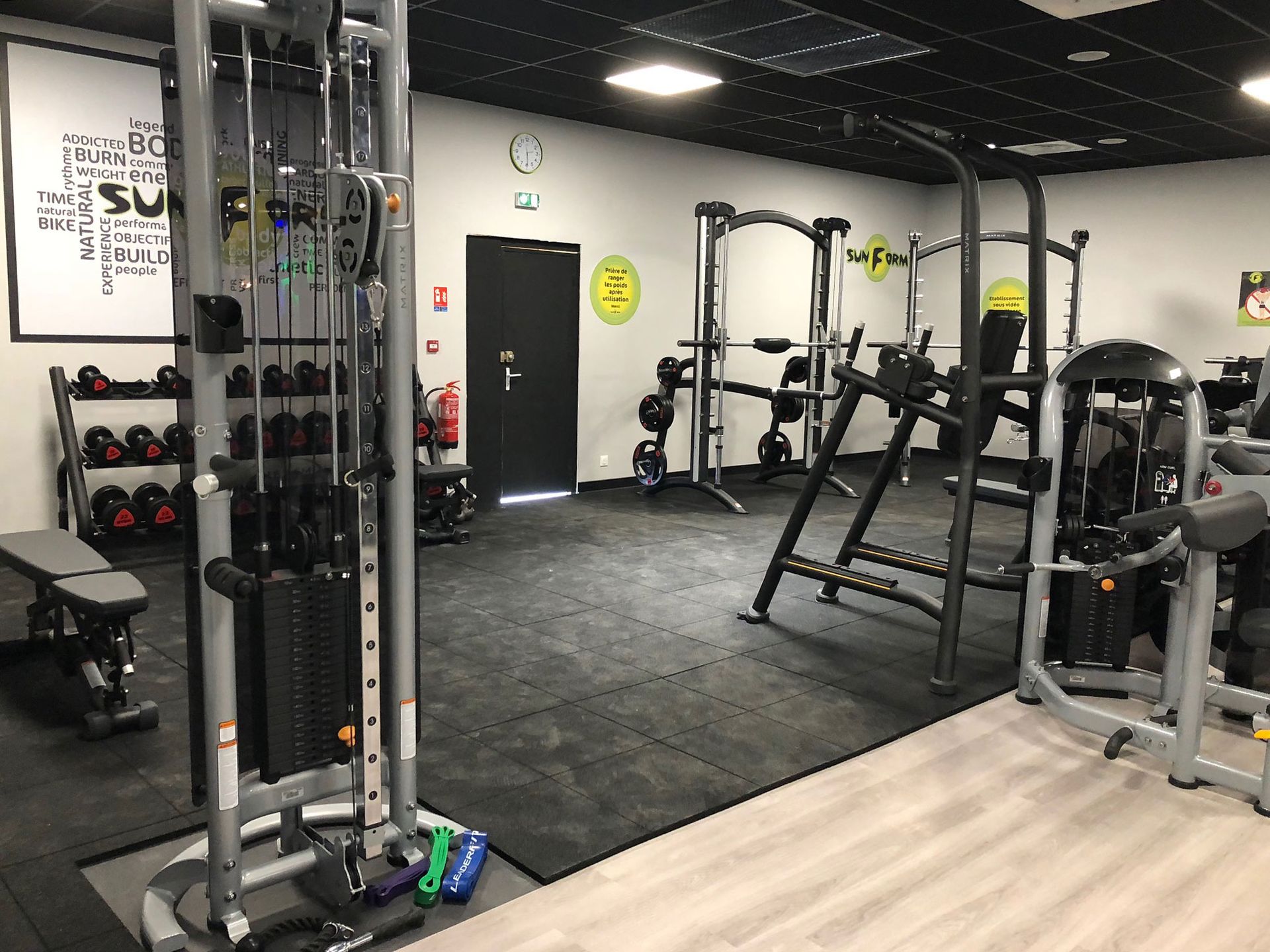 Intérieur d'une salle de sport avec machines de musculation, haltères et banc sur un sol en caoutchouc noir.