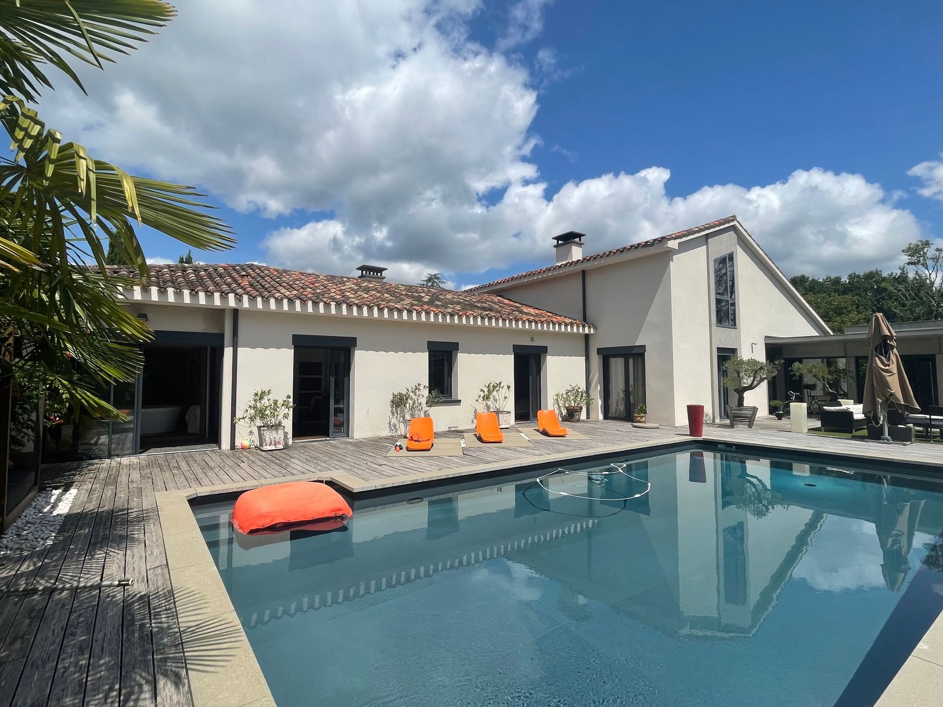 Piscine devant une maison aux couleurs claires, avec des volets foncés et des chaises orange sur une terrasse, sous un ciel bleu.