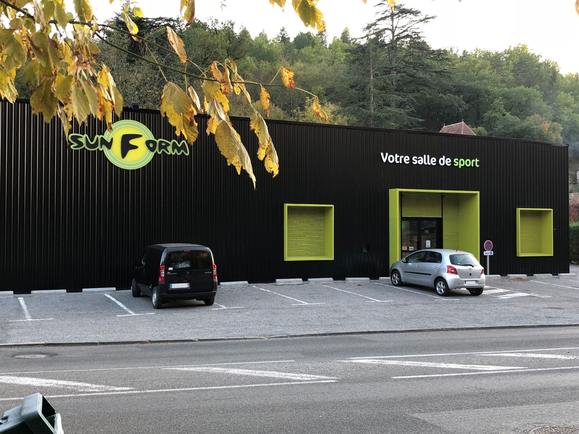 Bâtiment noir avec des touches de vert, logo « SUN FORM », voitures garées, arbres en arrière-plan.