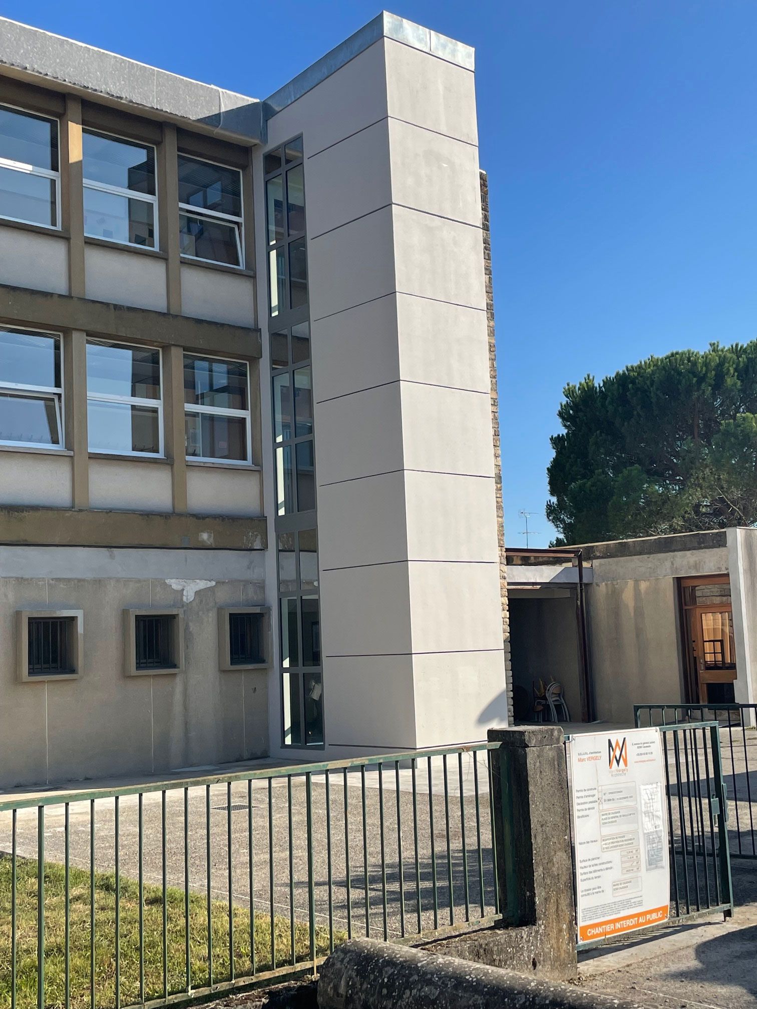 Vue extérieure d'un bâtiment scolaire avec une tour claire nouvellement construite ; journée ensoleillée.