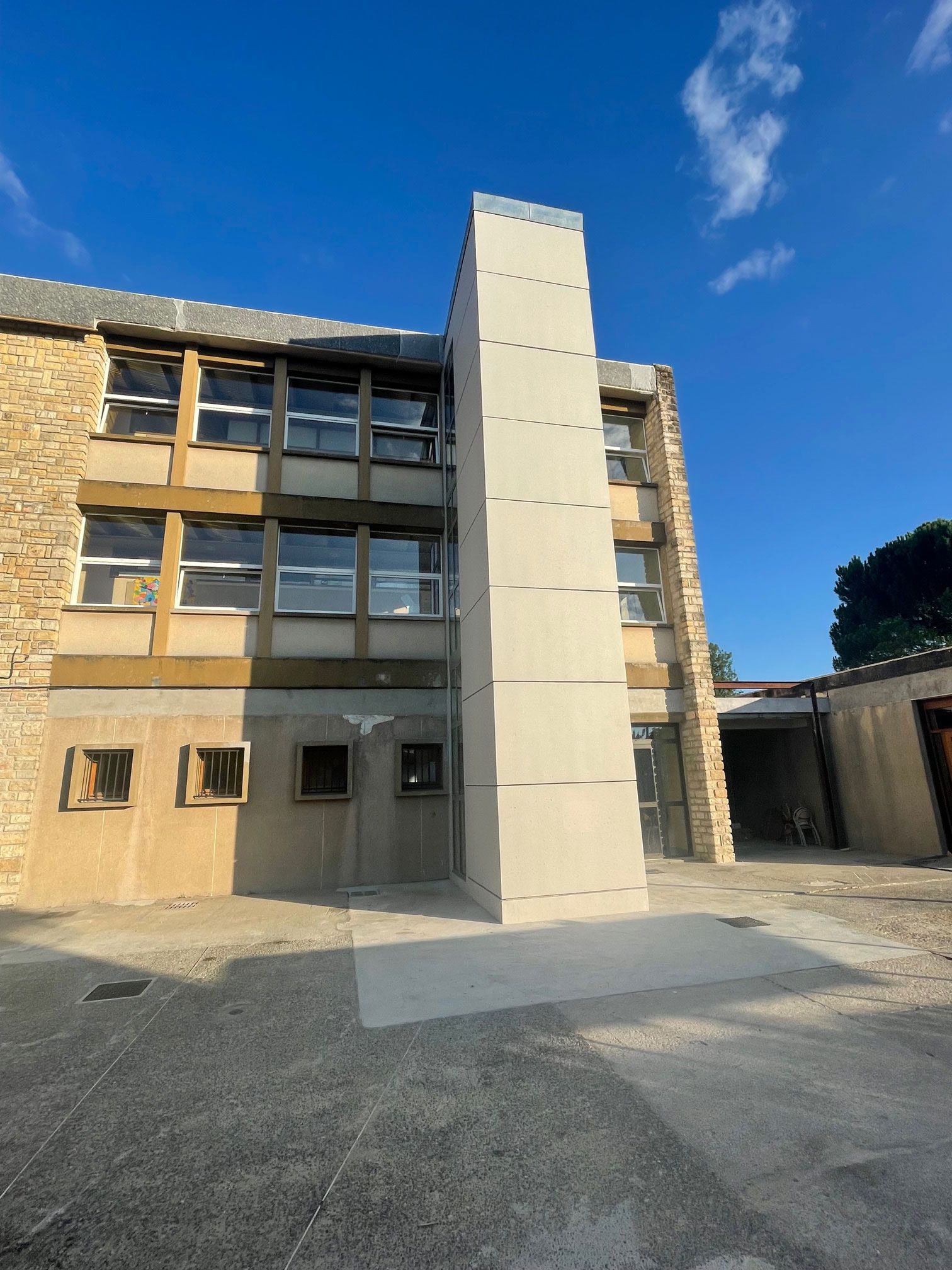 Vue extérieure d'un immeuble de plusieurs étages avec une haute structure en béton clair attenante. Ciel bleu au-dessus.