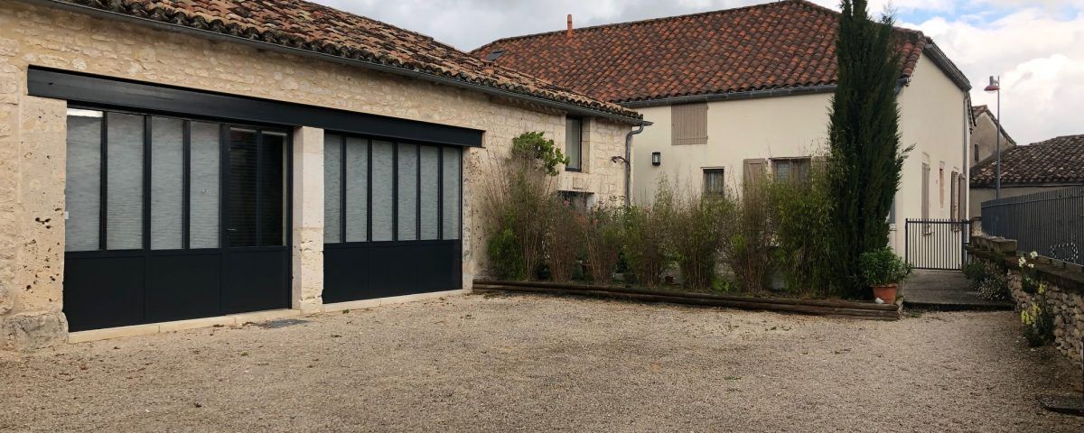 Une cour gravillonnée avec des bâtiments ; une porte-fenêtre à cadre sombre, un toit de tuiles rouges et une grande plante verte.