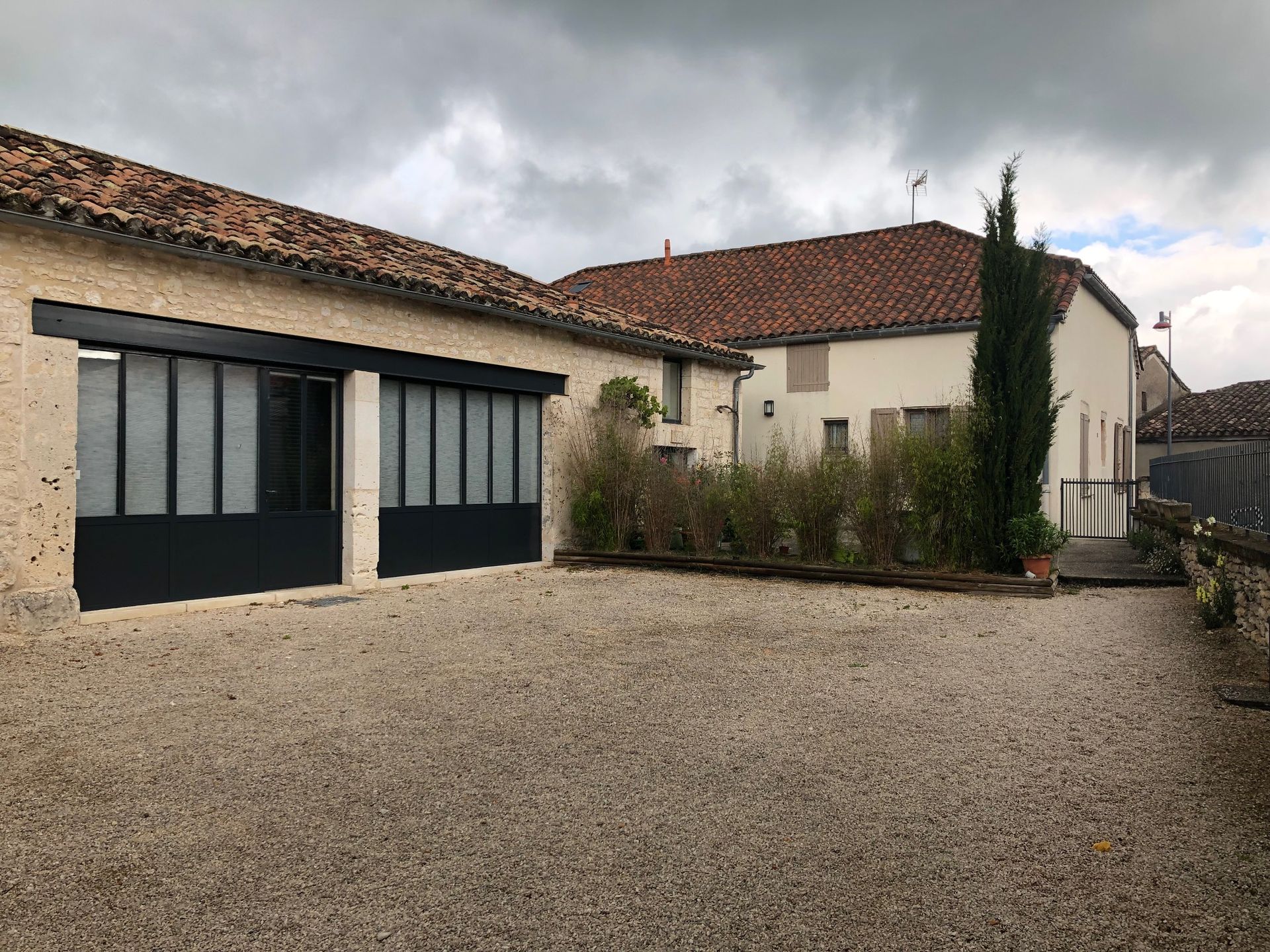 Cour intérieure gravillonnée avec des bâtiments beiges, des fenêtres à encadrement noir et un toit de tuiles en terre cuite sous un ciel nuageux.