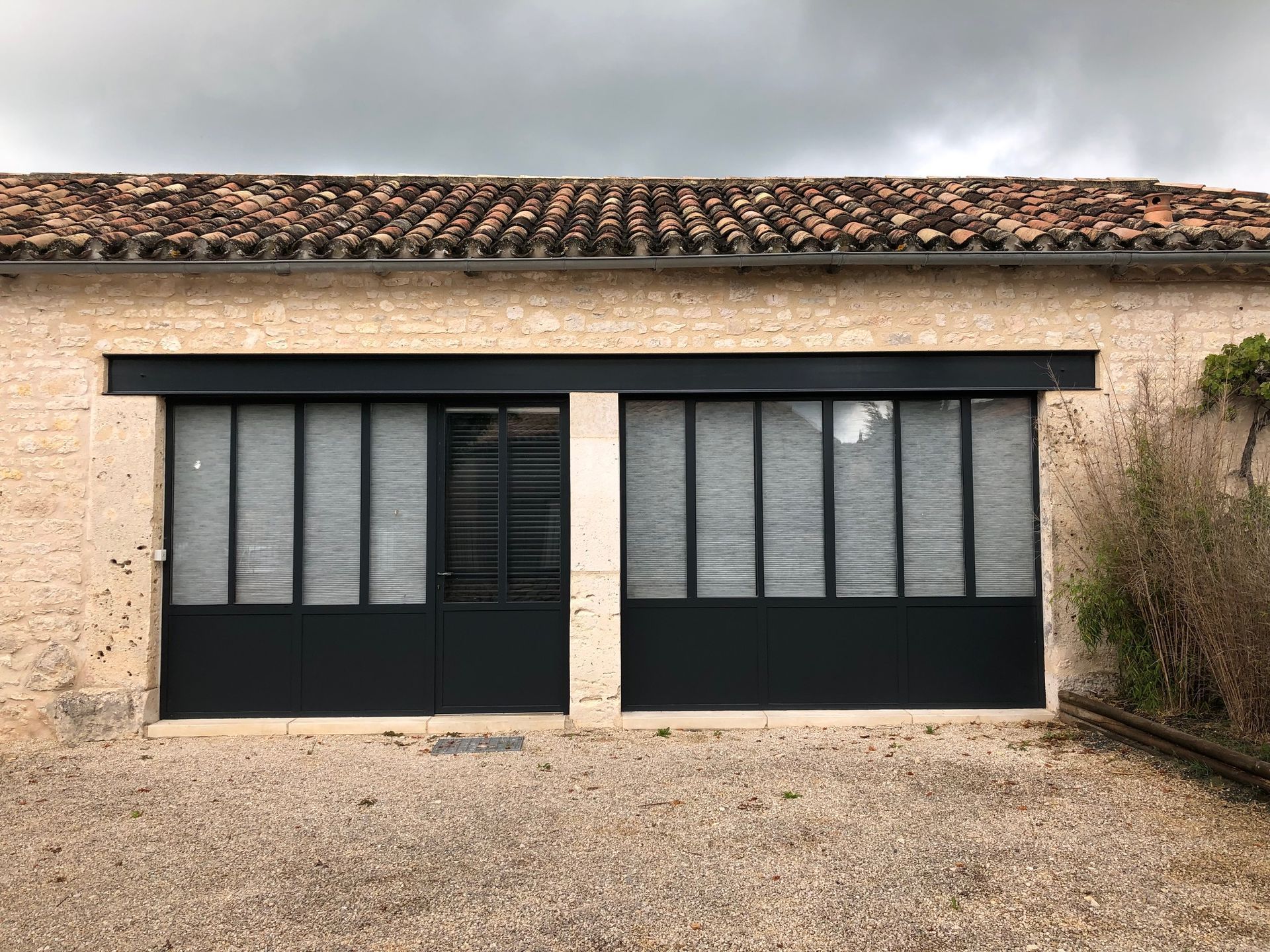 Extérieur d'un bâtiment en pierre beige avec des fenêtres à encadrement noir et une porte sous un toit de tuiles, ciel couvert.