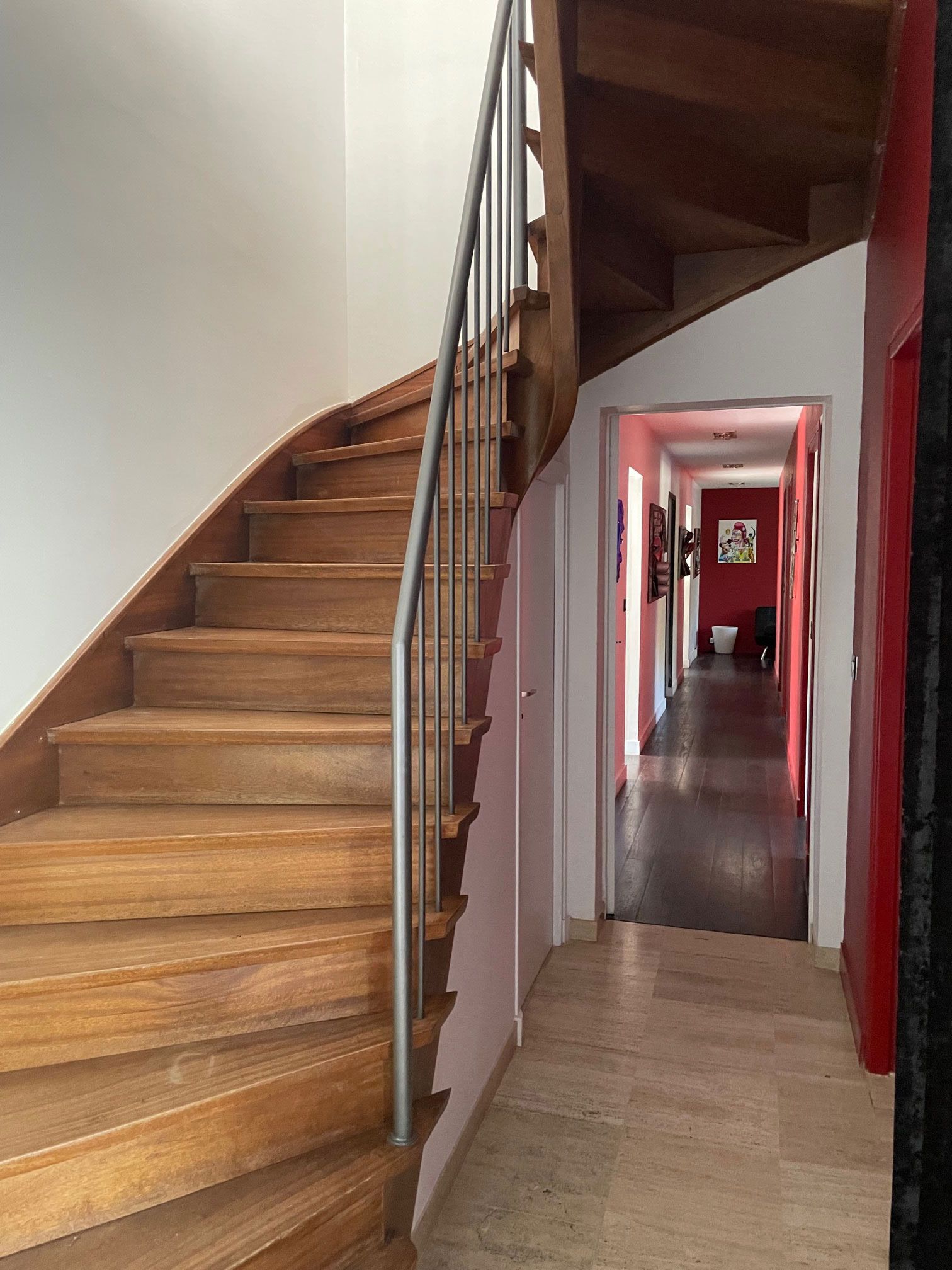 Un escalier en bois avec une rampe en métal mène à un couloir avec un mur d'accent rouge.