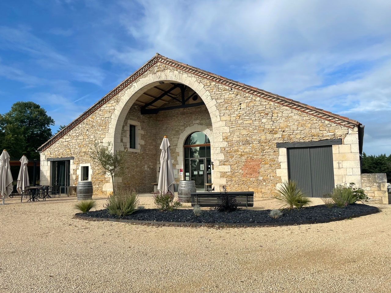 Bâtiment en pierre avec entrée voûtée et volets sombres ; petites plantes et gravier.