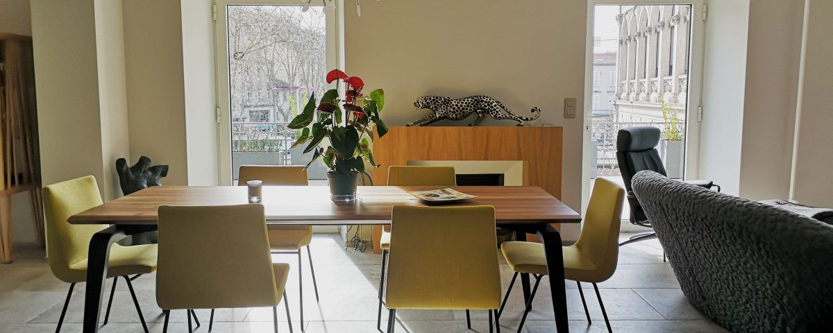 Salle à manger avec table en bois, chaises jaunes et fleurs rouges. Balcon en arrière-plan.