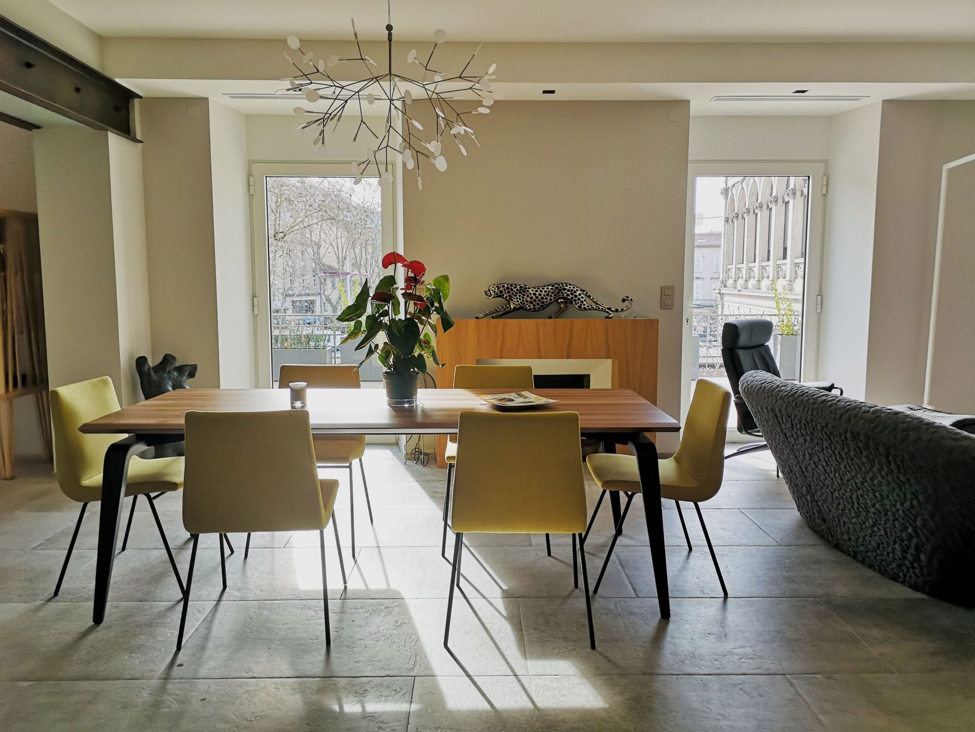 Salle à manger avec table en bois, chaises jaunes et lustre moderne. La lumière naturelle y pénètre abondamment.