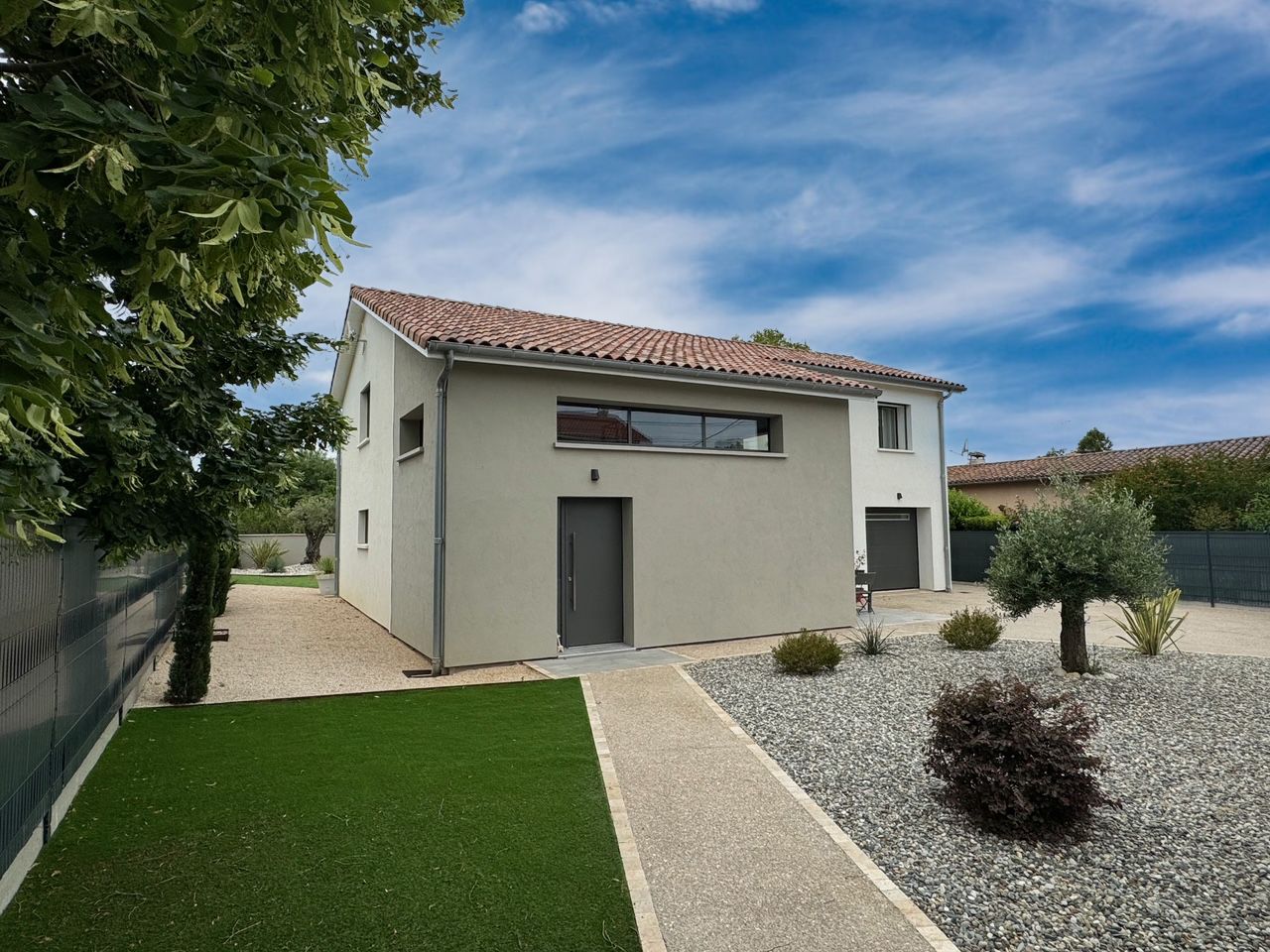 Maison à deux étages avec façade en stuc gris, toit de tuiles rouges et aménagement paysager en gravier sous un ciel bleu.