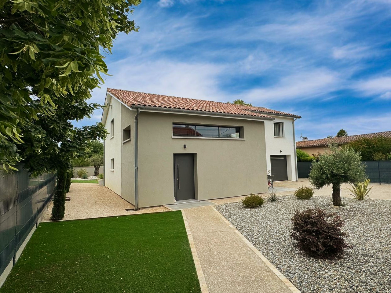 Maison à deux étages avec façade en stuc gris, toit de tuiles rouges et aménagement paysager en gravier sous un ciel bleu.