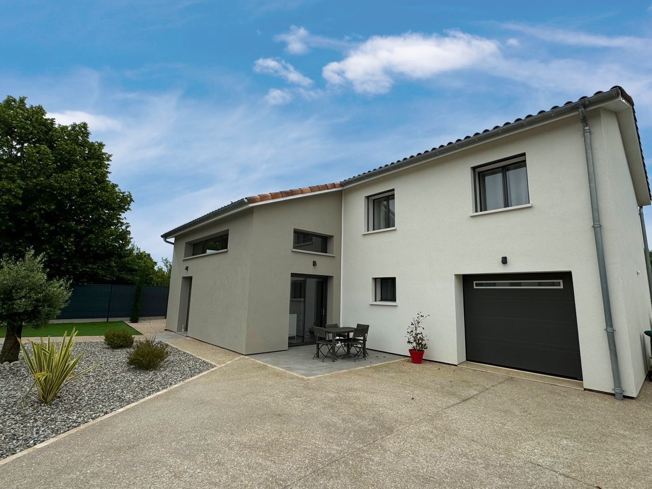 Maison moderne à deux étages avec façade grise, garage et allée en pierre sous un ciel bleu nuageux.