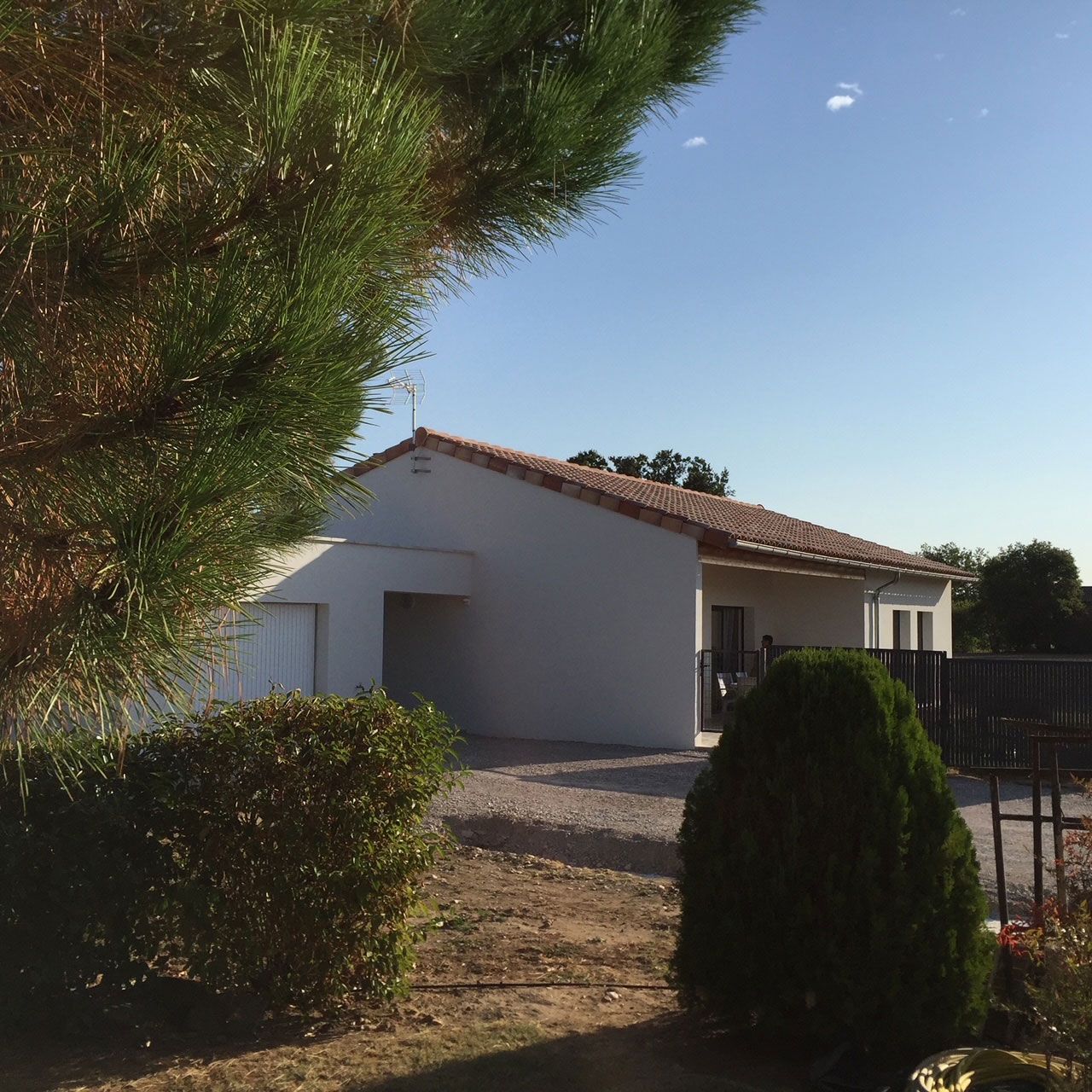 Maison blanche au toit de tuiles rouges, allée de gravier et verdure environnante sous un ciel bleu.