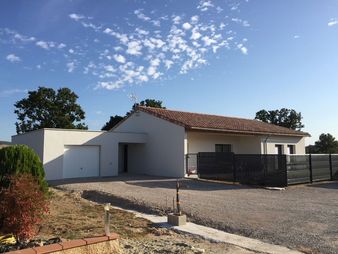 Maison blanche moderne avec un toit de tuiles en terre cuite, une allée de gravier et un ciel bleu nuageux.