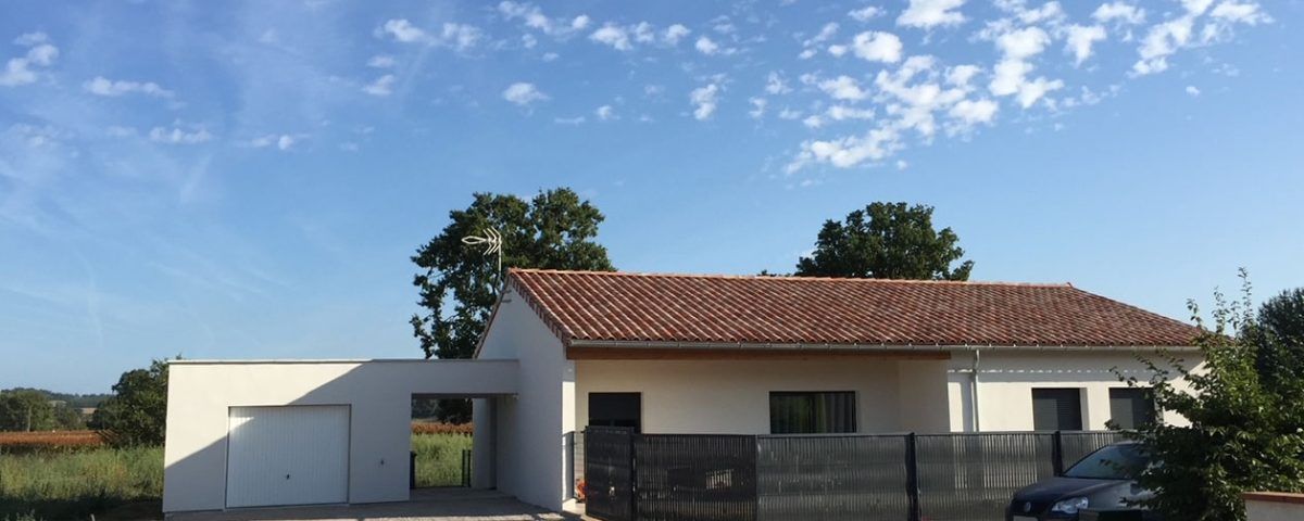 Une maison au toit de tuiles rouges, aux murs blancs et avec un garage indépendant, se détachant sur un ciel bleu nuageux.