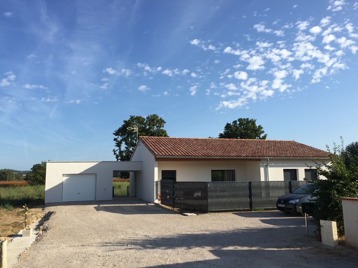 Maison blanche au toit de tuiles rouges, abri voiture, clôture noire, allée de gravier, sous un ciel bleu nuageux.