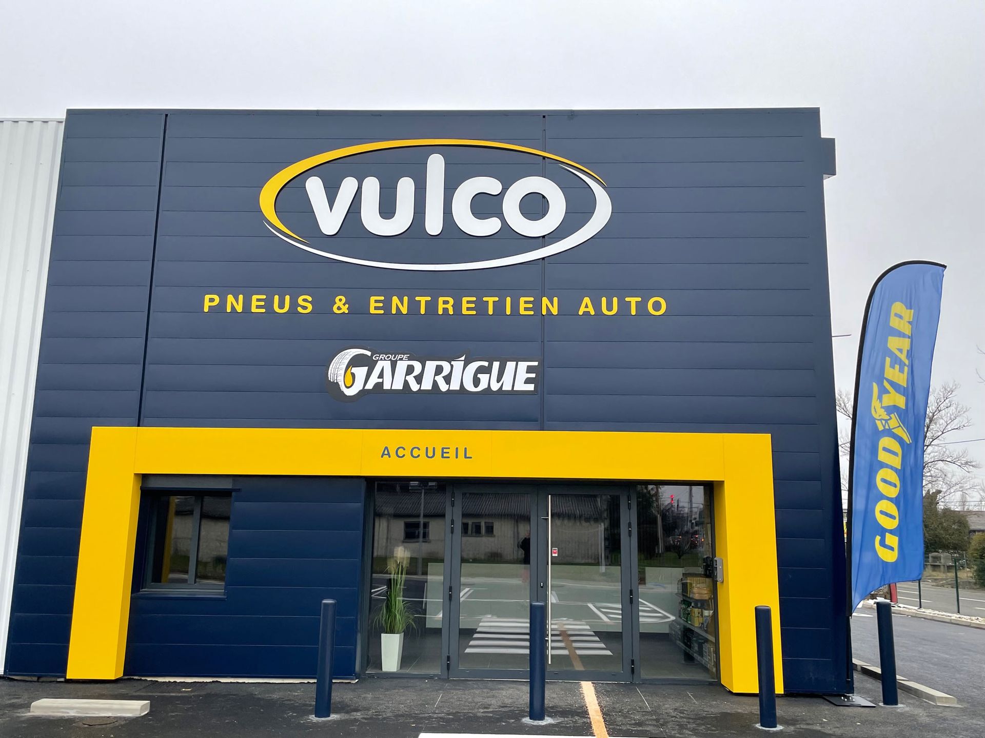 Magasin de pneus Vulco avec une façade jaune et bleu marine et un drapeau Goodyear.