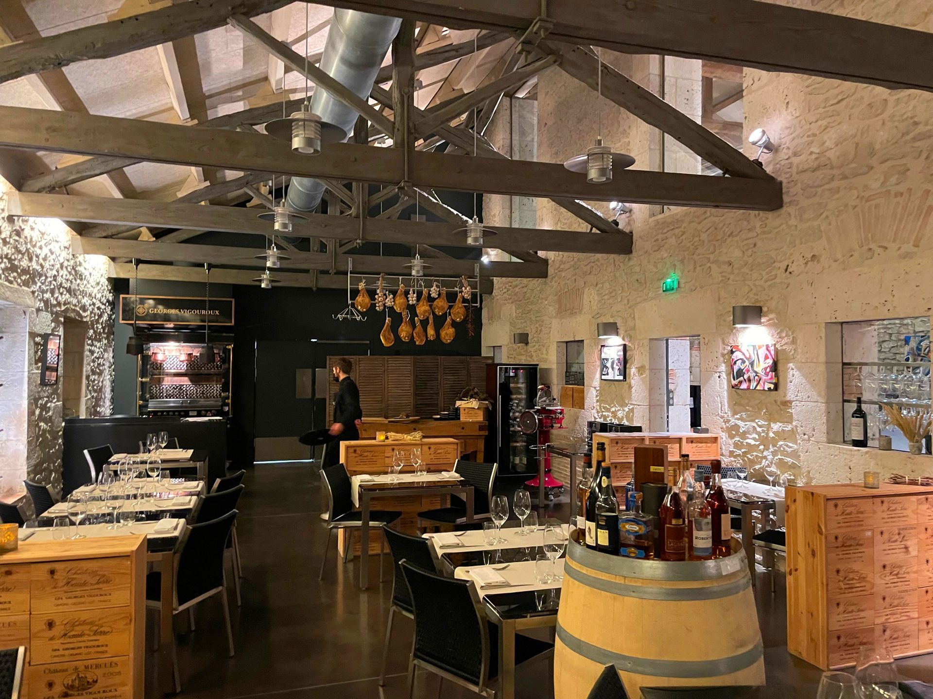 Intérieur d'un restaurant avec tables en bois et poutres apparentes. Une personne se tient près d'un comptoir où sont disposées des bouteilles.