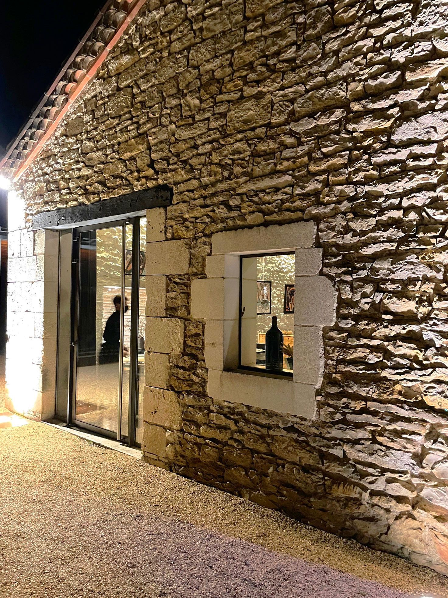 Bâtiment en pierre avec porte et fenêtre vitrées la nuit, chemin de gravier éclairé.