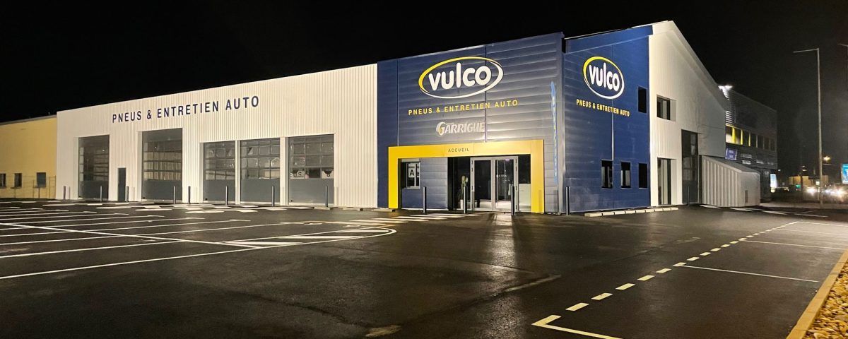 Extérieur d'un garage Wilco, de nuit. Le bâtiment est bleu et blanc avec des touches de jaune. Il pleut au sol.